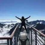 woman standing on a guardrail overlooking clouds and mountaintops.